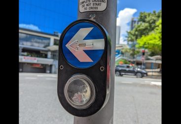 A close up of street pedestrian button with the arrow facing left. The button is black, with a round stainless steel circle button and a blue circle with a silver stainless steel arrow.