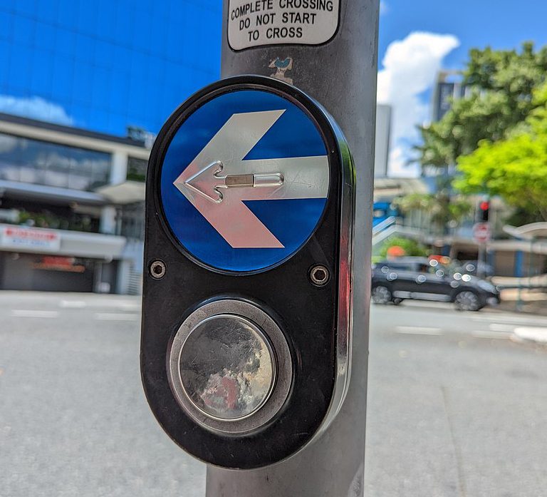 A close up of street pedestrian button with the arrow facing left. The button is black, with a round stainless steel circle button and a blue circle with a silver stainless steel arrow.